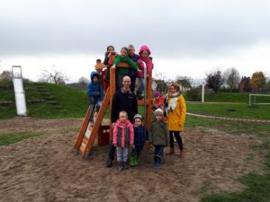 Eine Gruppe von Kindern und Erwachsenen steht auf einem Spielplatz neben einer Holzrutsche und einem Klettergerüst.