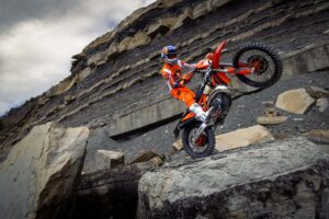 Ein Motorradfahrer zeigt seine Fähigkeiten auf einer KTM beim Fahren über einen großen Felsen in einer steinigen Umgebung.
