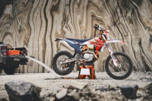 Ein KTM-Motorrad steht auf einem Ständer vor einer beeindruckenden, geschichteten Felswand, während ein Pickup im Hintergrund zu sehen ist.