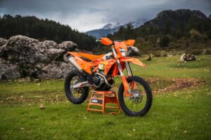 Eine orangefarbene KTM Enduro-Motorrad steht auf einem Ständer in einer grünen Landschaft mit Felsen und Bäumen im Hintergrund.