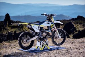Husqvarna Enduro-Motorrad auf einer Werkstattmatte in einer malerischen Landschaft mit Bergen im Hintergrund.